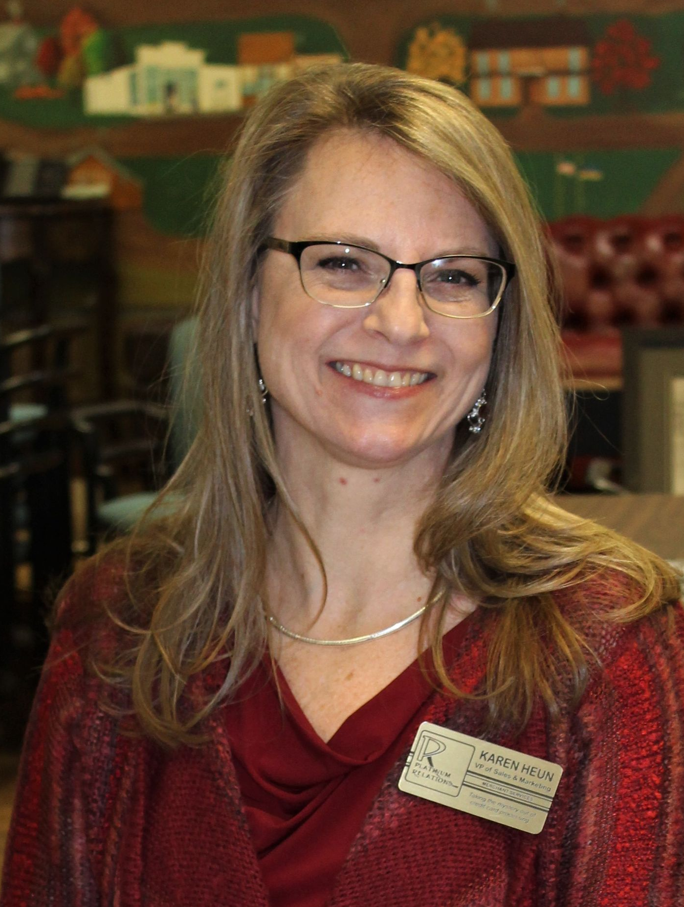 Woman with glasses smiles, wearing a red shirt, indoors, in front of blurred background.
