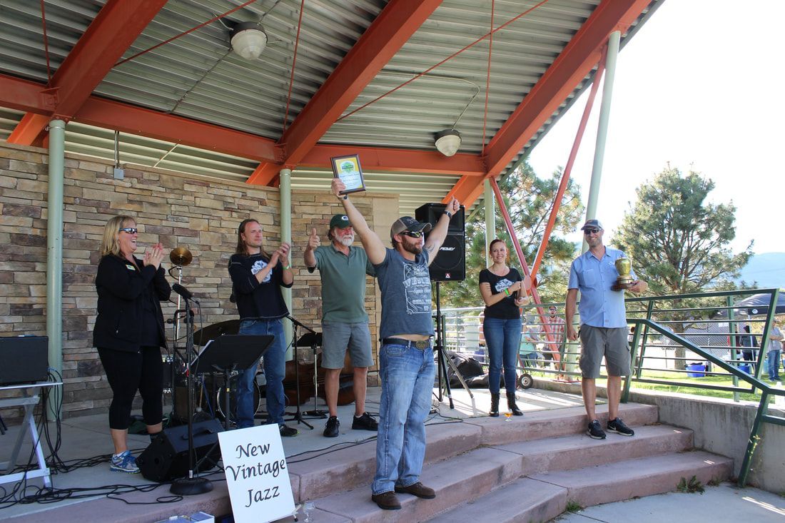 Group celebrates on outdoor stage; man holding trophy; others clap, trees in background.