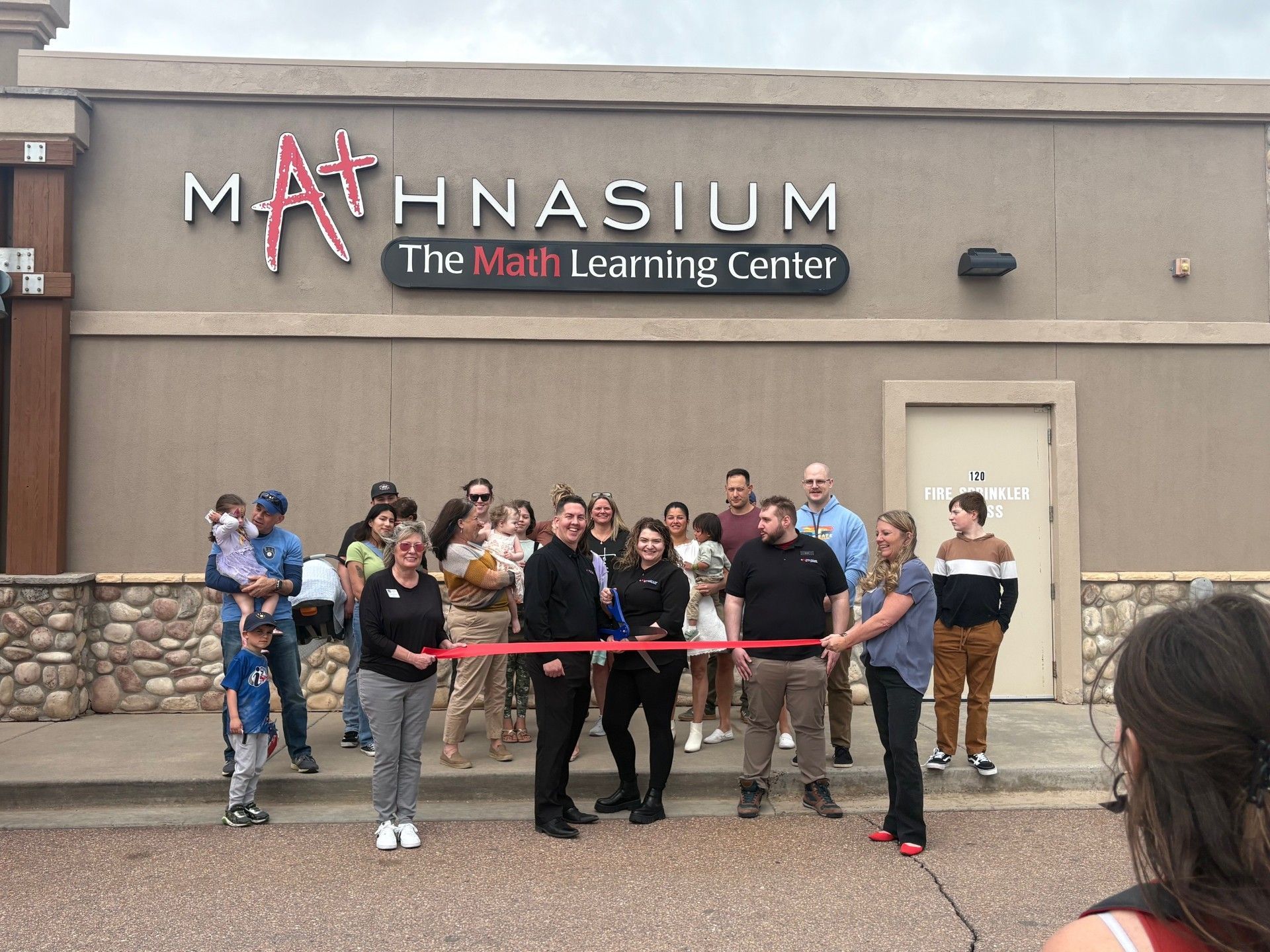 Ribbon-cutting ceremony outside Mathnasium learning center; group of people, red ribbon, brown building.