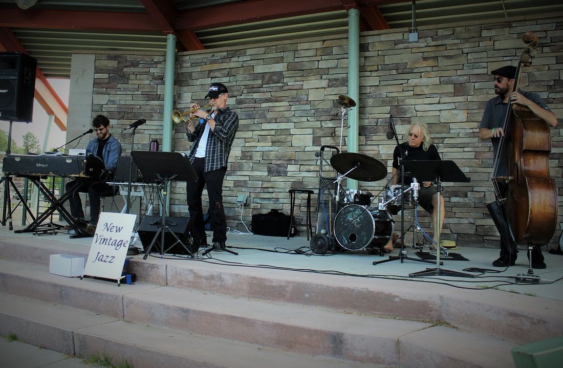 Jazz band performing outdoors on a stage. Musicians include trumpet, keyboard, drums, and bass.
