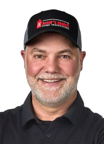 Man with a gray beard and ball cap, wearing a plaid shirt, smiling, against a blue background.