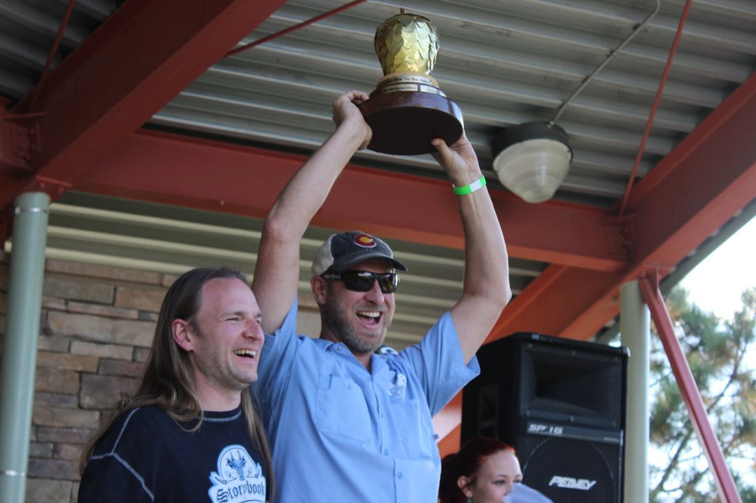 Man holds trophy aloft, smiling, on a stage under a roof with another man; outdoor setting.