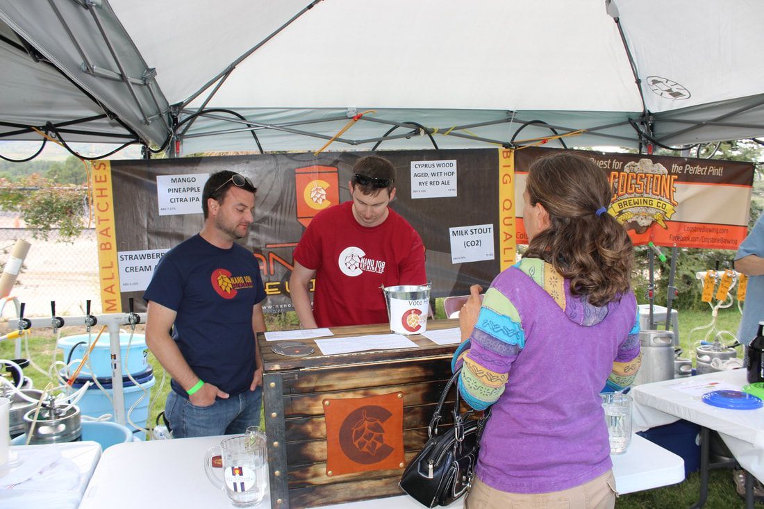 Two men in a brewery stand at a booth, serving beer to a woman with a colorful sweater.