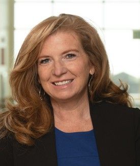 Woman with reddish-blonde hair smiles at the camera; wearing a blue top and black blazer.