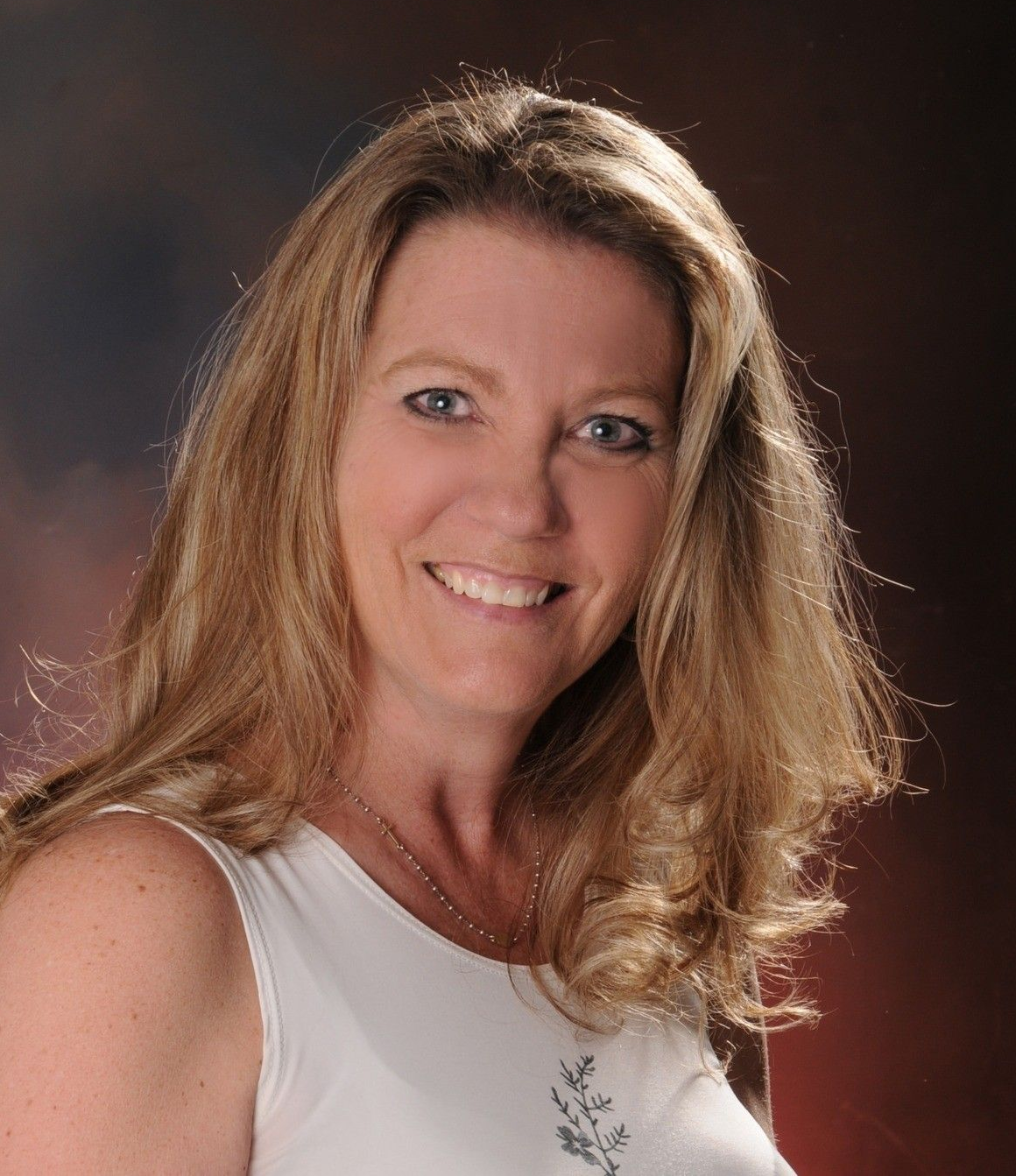 Woman with blonde hair smiling, wearing a white tank top, against a blurred background.