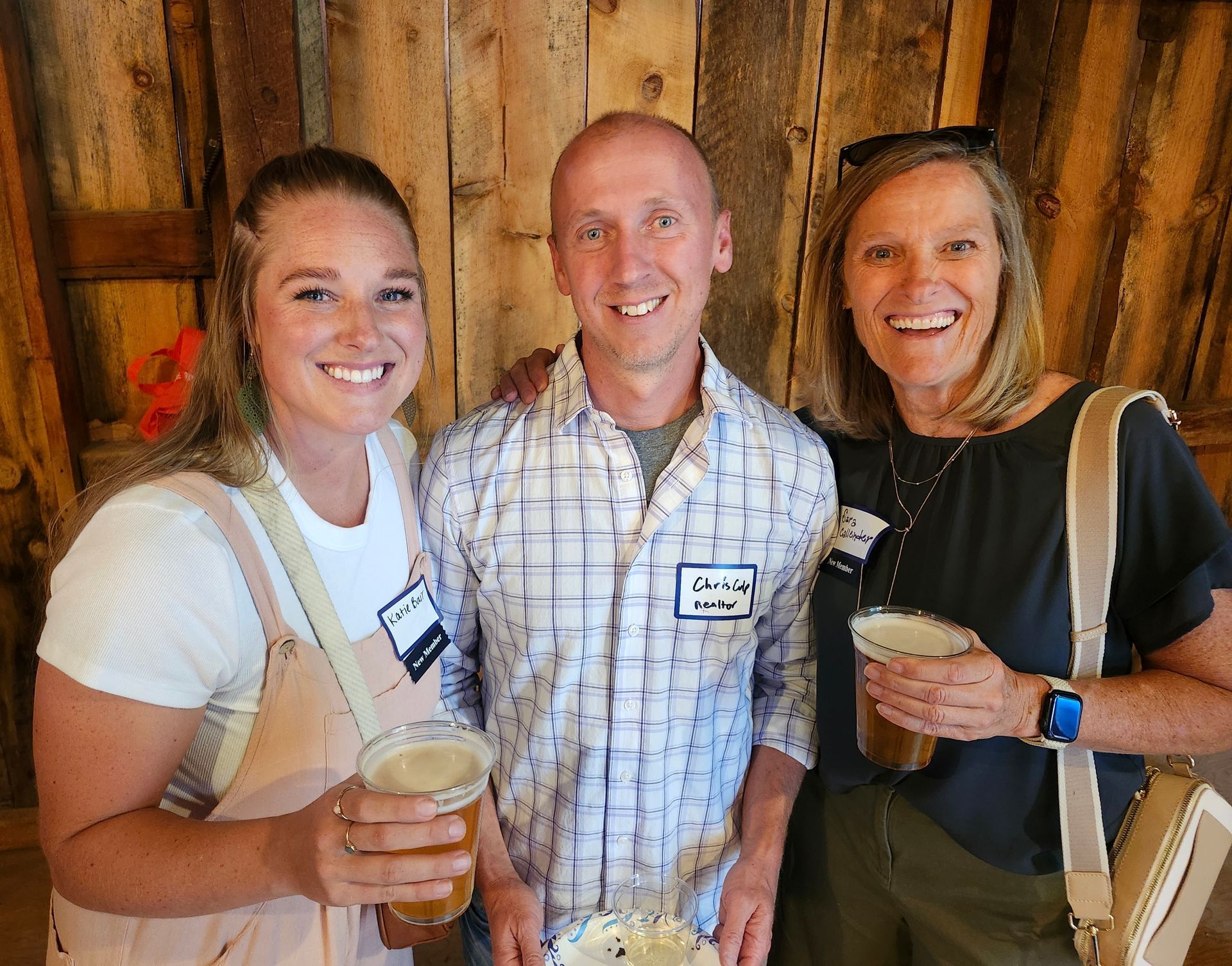 Three people smiling, holding drinks, in a wooden interior. One man has a hand on his shoulder.
