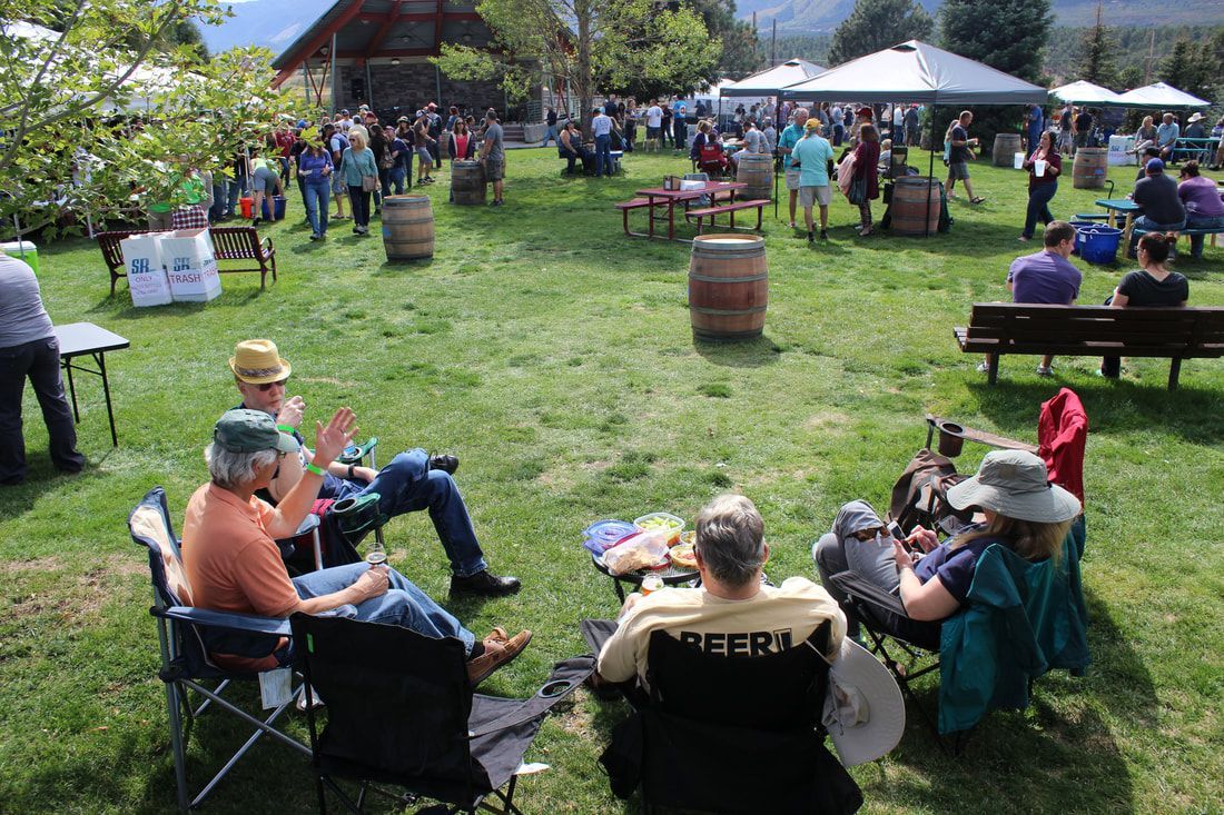 People enjoying an outdoor event in a grassy area, with wine barrels, tents, and a building in the background.