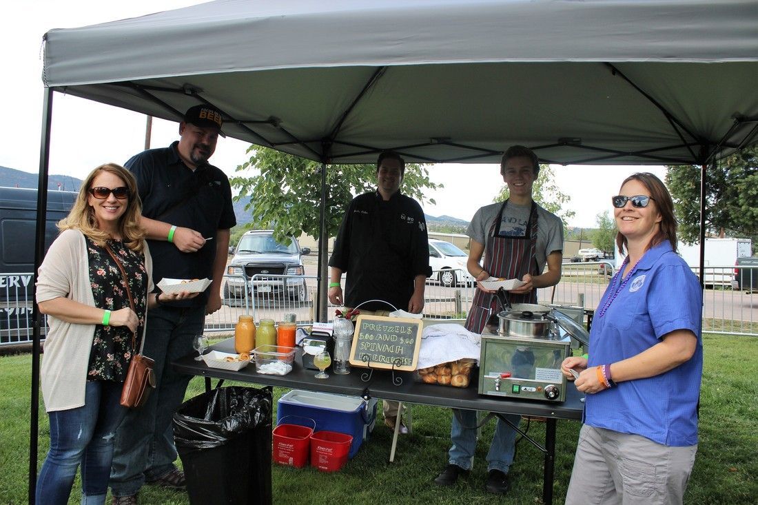 People at a food vendor booth under a canopy, with food and drinks on a table.
