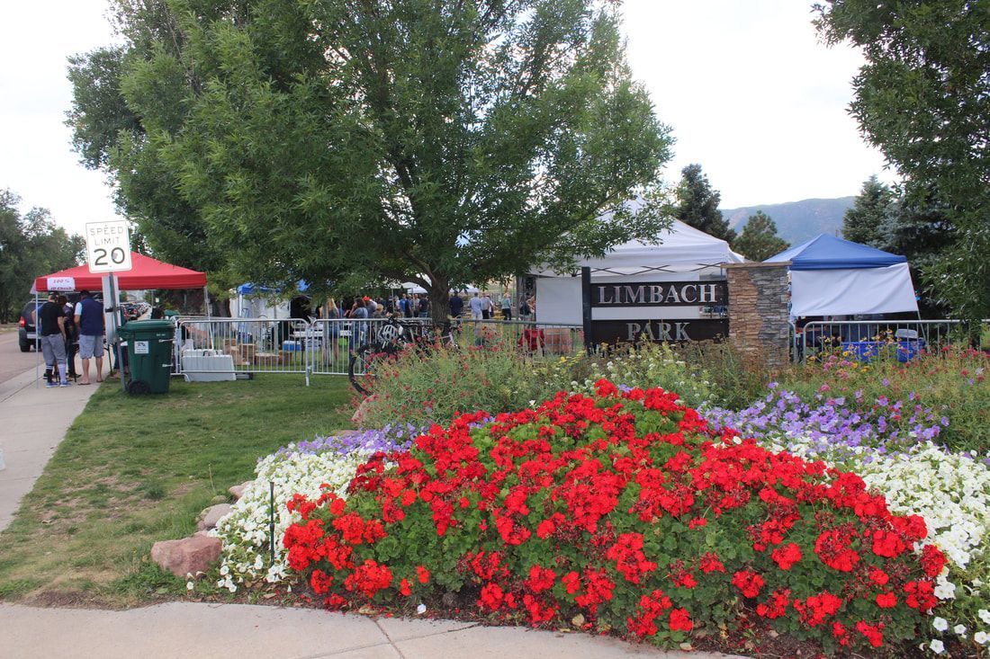 People at a park market with red and white flower beds in front of an entrance sign.