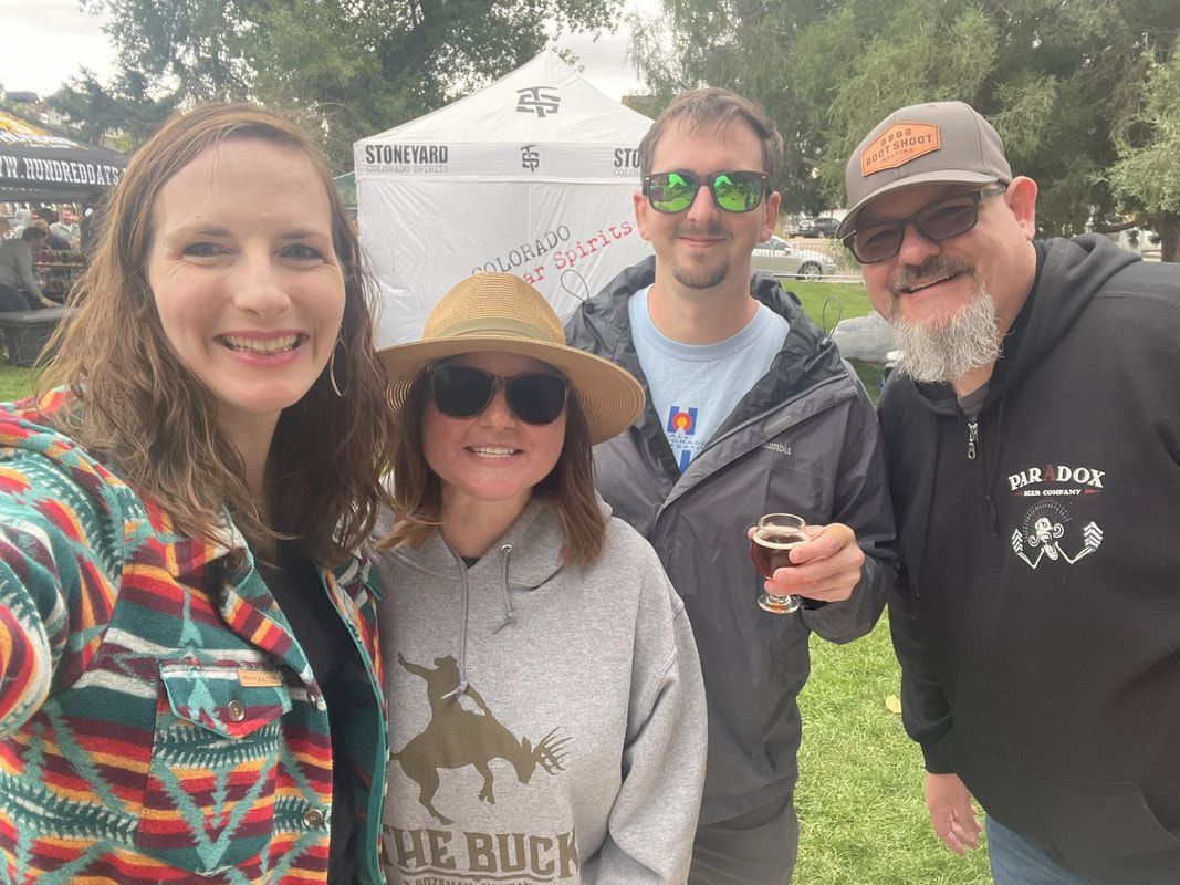 Four people smiling at an outdoor event. Two women, two men, near a tent, holding a beer.