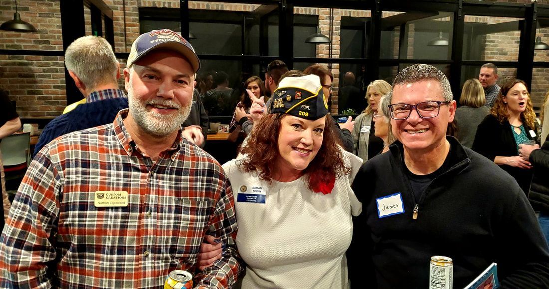 Three people smiling, arm around each other, in a crowded room. Woman wears a veteran hat.