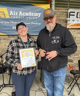 Woman in plaid, holding award, smiles with man, under a banner for Air Academy Credit Union at outdoor event.