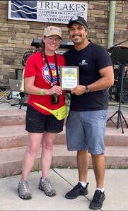 Two people outdoors holding an award. Man in black shirt, woman in red shirt, near Tri-Lakes Visitor Center sign.