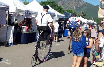 People on tall bikes at an outdoor festival. One man in a beret rides near booths. Others watch.