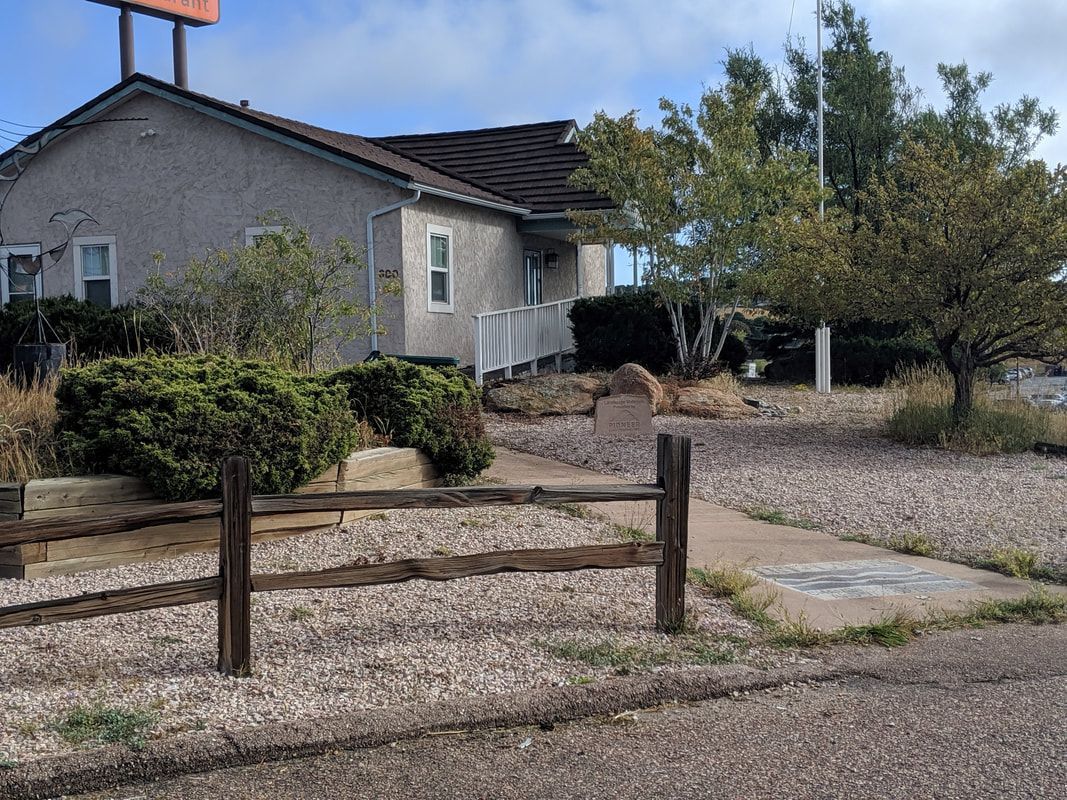 A small, stucco building with a brown roof, gravel yard, and a wooden fence in front.