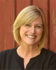 Woman with blonde hair smiles in front of a red wooden wall, wearing a black shirt.