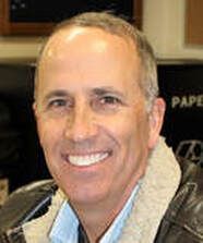 Man with short gray hair smiles, wearing a blue shirt and a brown jacket with a gray collar.