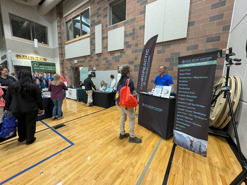 Health fair. A man at a chiropractic booth with a sign; people browse other booths in a gym.