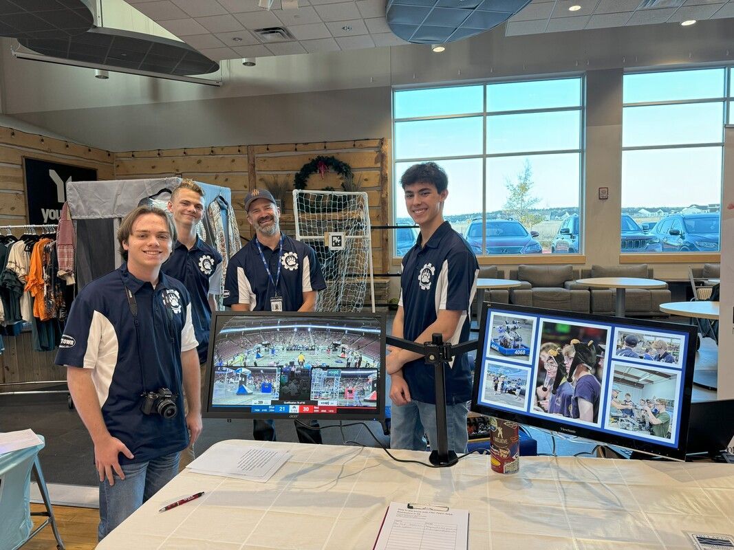Four men in matching blue shirts stand behind a table with monitors. They appear to be at an event.