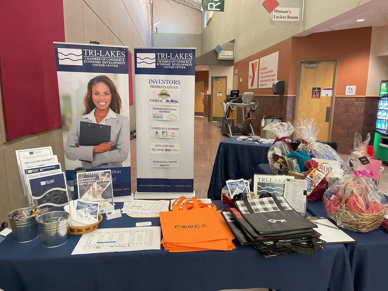 A table at an event with promotional materials for Tri-Lakes. Banners, baskets, and brochures are displayed.