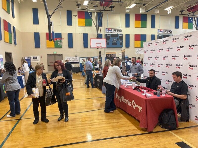 People at a business expo in a gymnasium. Attendees greet exhibitors at booths with red table cloths.