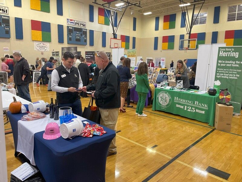 People interacting at a business fair in a gymnasium. Tables, signage, products, and attendees are visible.