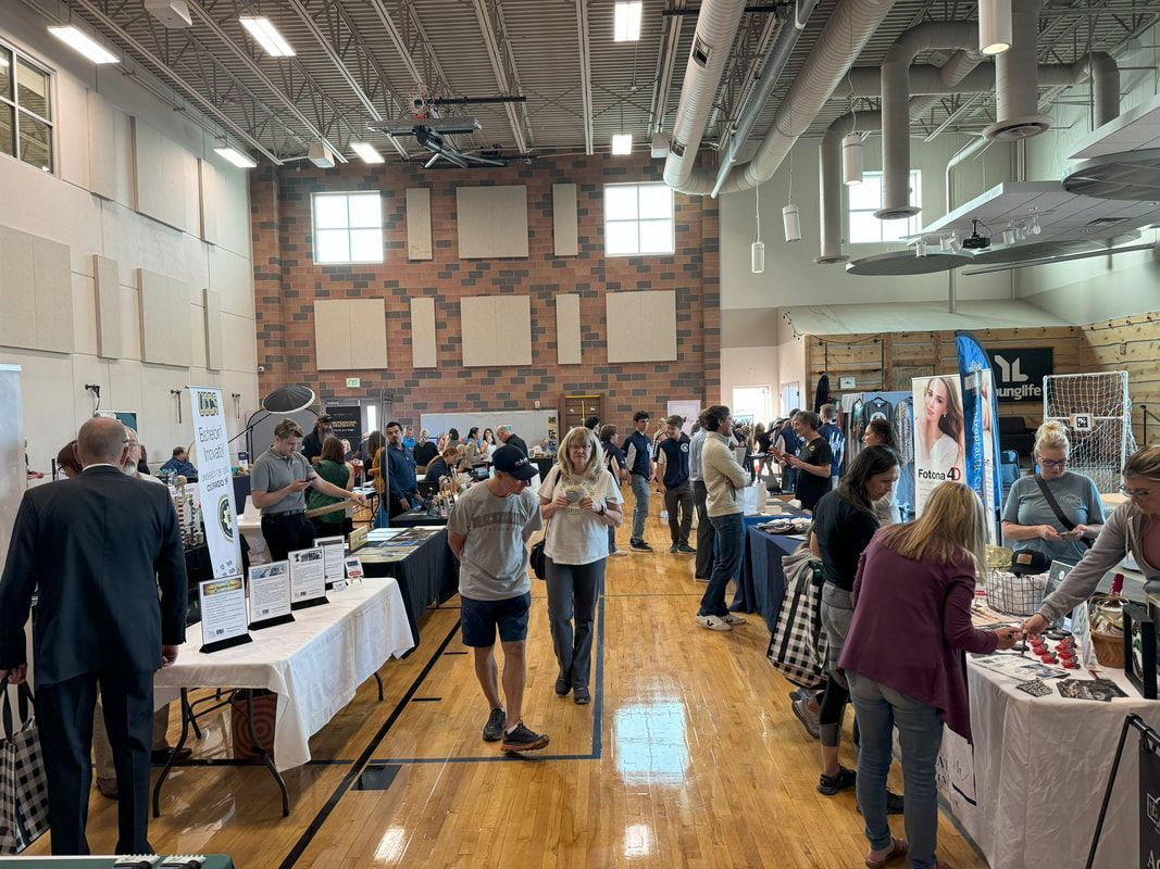 People browse vendor booths at an event in a gymnasium with wood floors and high ceilings.