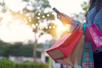 Woman holding shopping bags, looking at phone outdoors, sunlight.