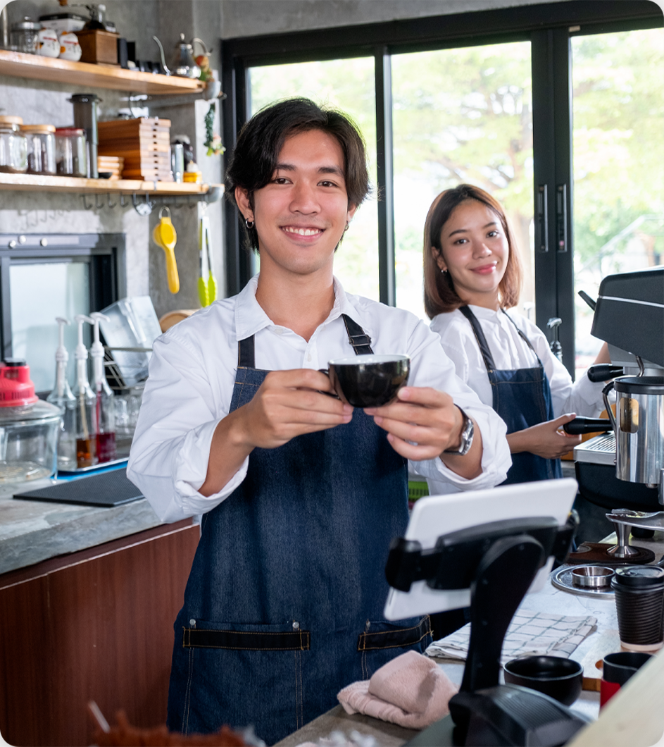 Two baristas smiling, holding coffee cups behind a cafe counter.