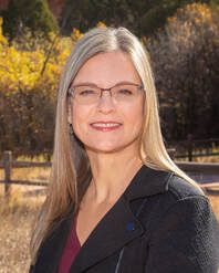 Blonde woman with glasses smiles outdoors. She wears a black blazer, red shirt, and poses in front of a blurred fall landscape.