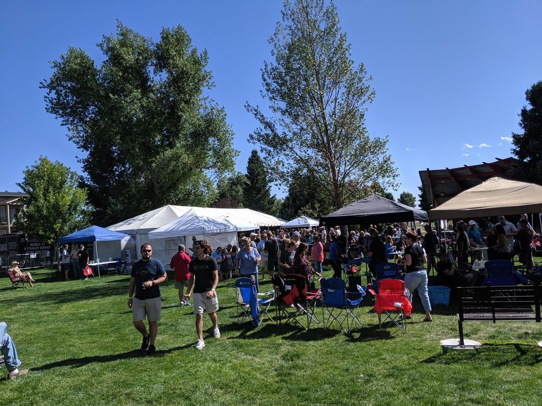 People gather on a grassy field under a bright blue sky, some in chairs, near tents and trees.