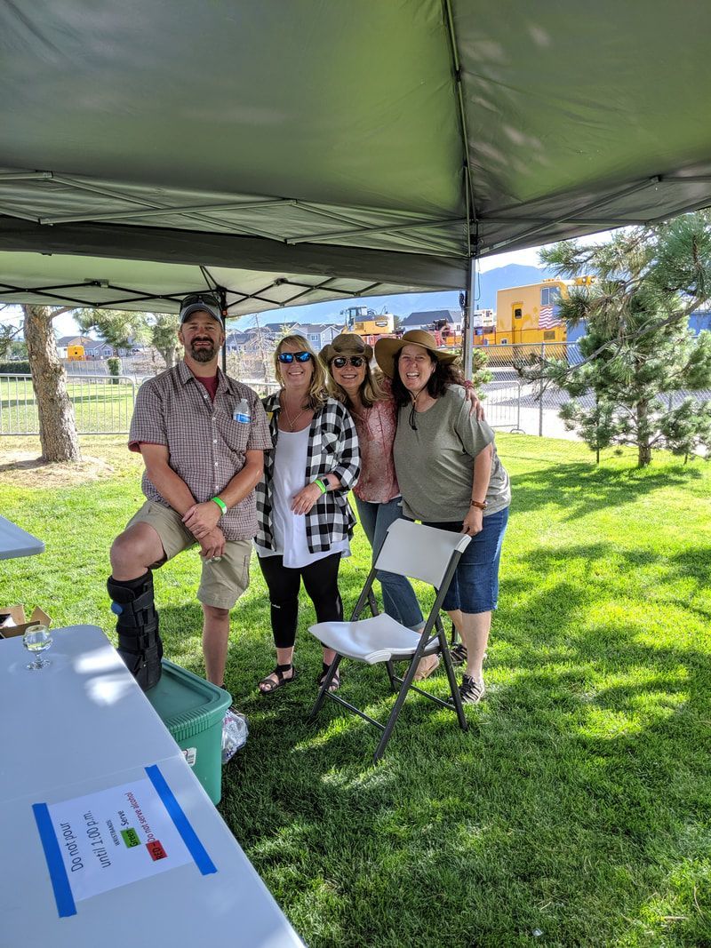Group of people under a tent outdoors. Man with leg brace, three women smile. Green grass, table with sign.
