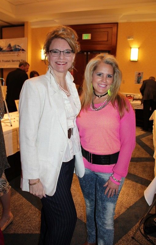 Two women smiling, one in a white blazer and patterned pants, the other in pink sweater and acid-wash jeans.