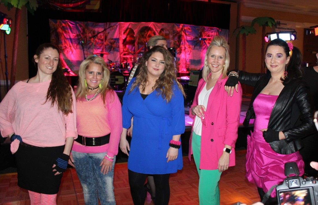 Five women in 80s-themed outfits at a party. Bright pink and blue clothing, posing. Stage backdrop visible.