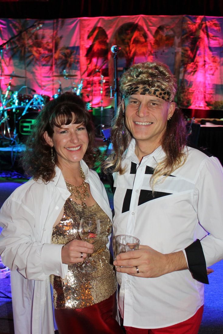 Woman and man at a party, the man wearing a mullet wig, the woman wearing a sequin top, both smiling.