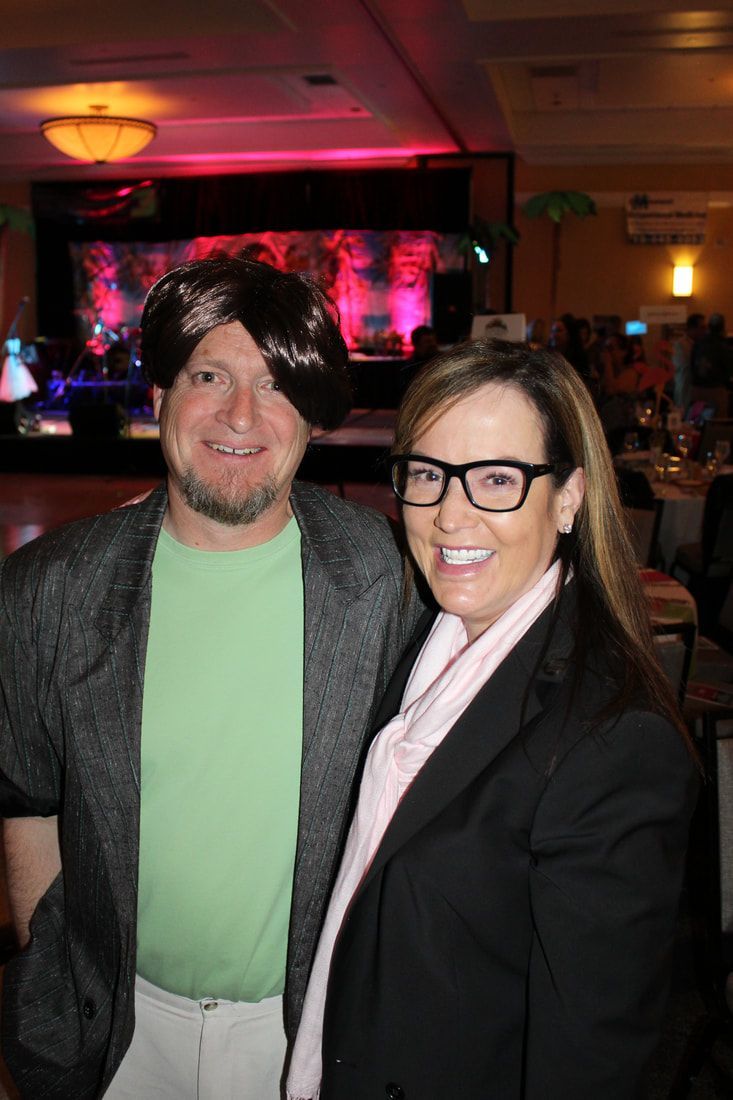 Man in wig and woman in blazer smile at event with stage backdrop.