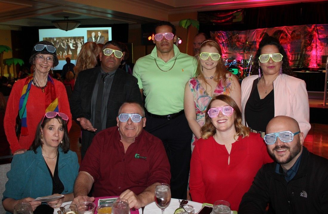 Group of people wearing neon-rimmed glasses, posing at a party with decorations and colorful lighting.