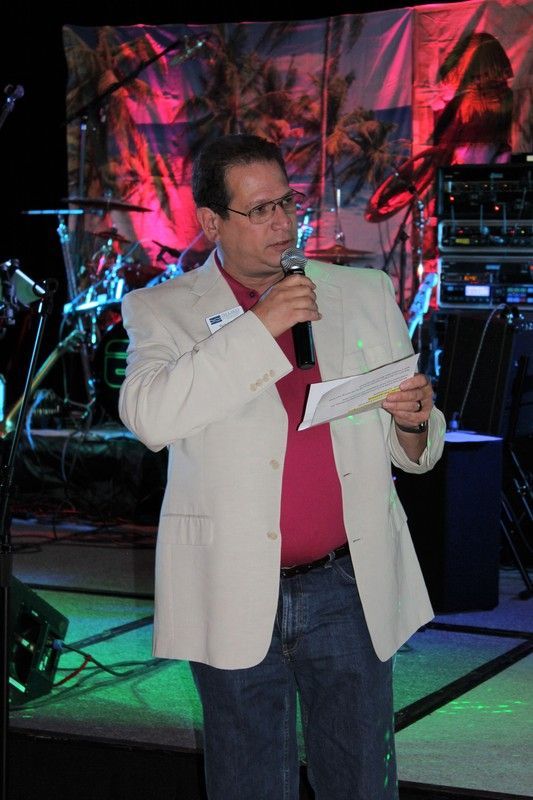Man in blazer speaks into microphone, holding paper, onstage with band equipment.