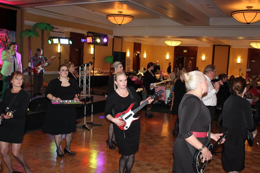 Band playing at an event. Several women in black dresses play instruments, while others dance in a brightly lit room.