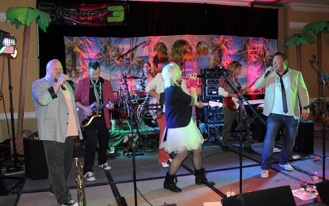 Band performing on stage, tropical theme with palm trees, bright lights, and several musicians with mics.