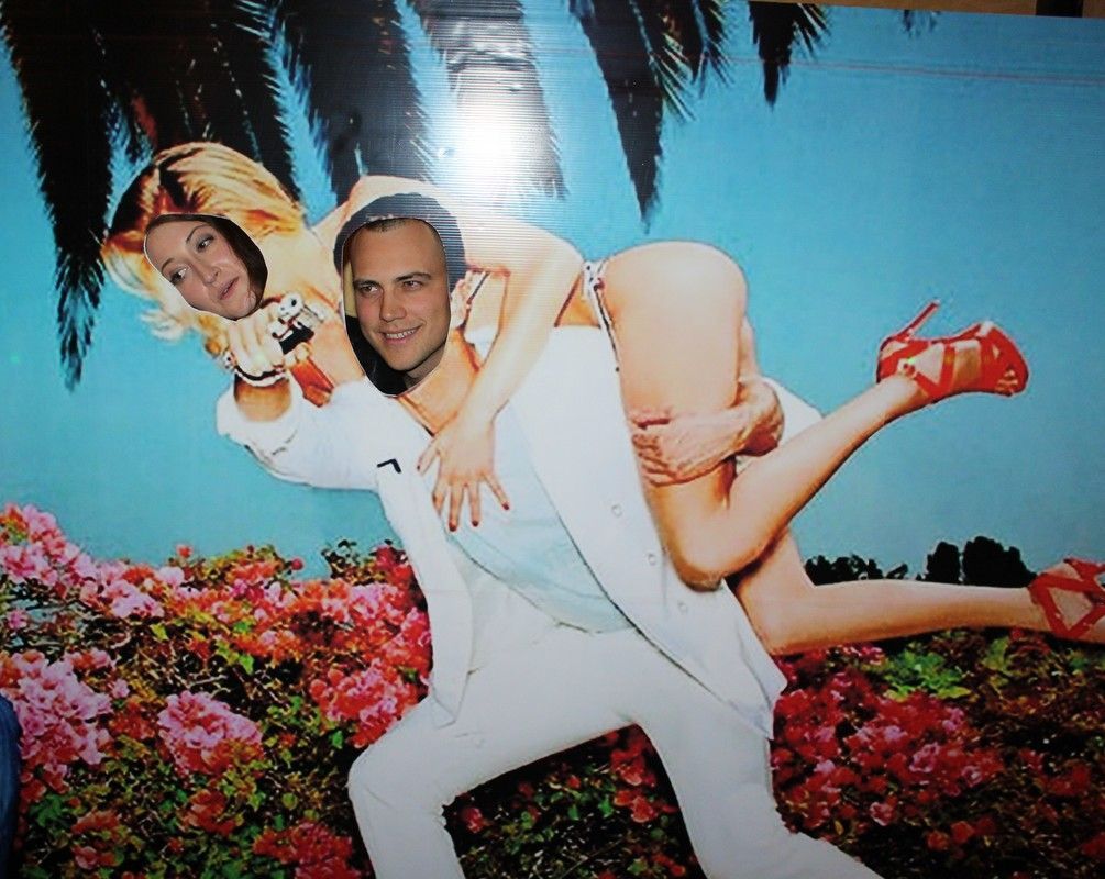Man posing in photo cutout, holding woman on back against a floral, tropical background.