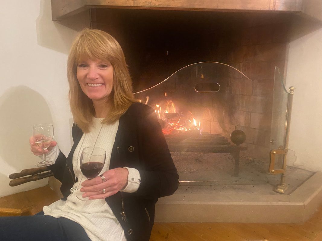 Woman with blonde hair smiling, holding wine, sitting by a lit fireplace.