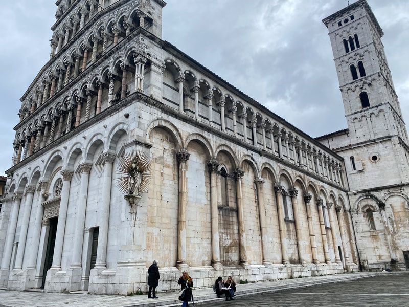 Exterior of a light-colored Italian church with ornate facade and tall bell tower under cloudy sky. People stand near the building.