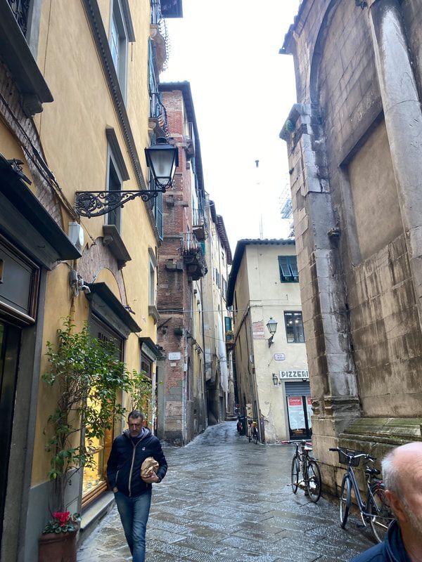 Narrow cobblestone street in Lucca, Italy, flanked by tall, colorful buildings. A man walks towards the viewer.