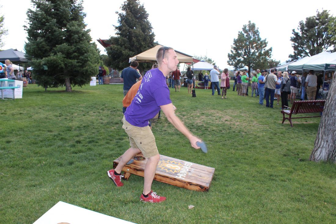 Man in purple shirt throws a bean bag at a cornhole board on a grassy lawn; a crowd watches.