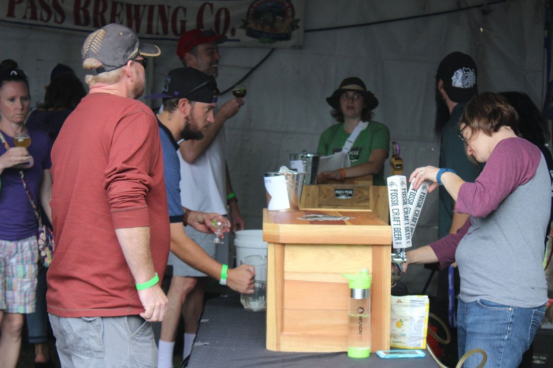 People at a beer tasting booth; a woman pours beer, others wait, logo in background.