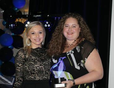 Two women smiling, one holding a trophy. Both dressed up, at an event with blue and black decor.