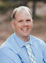 Smiling man wearing a light blue shirt and patterned tie outdoors.