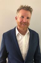 Man with red beard, wearing a blue blazer and white shirt, smiling in front of a light-colored wall.