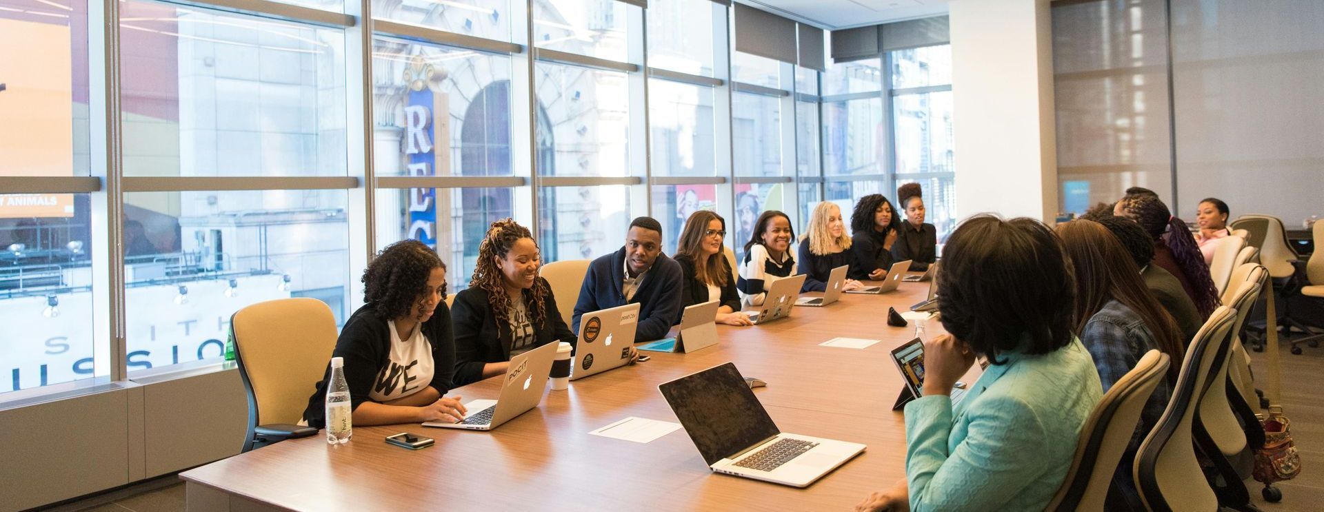 A diverse group of people sitting at a long table in a brightly lit conference room, working on laptops.